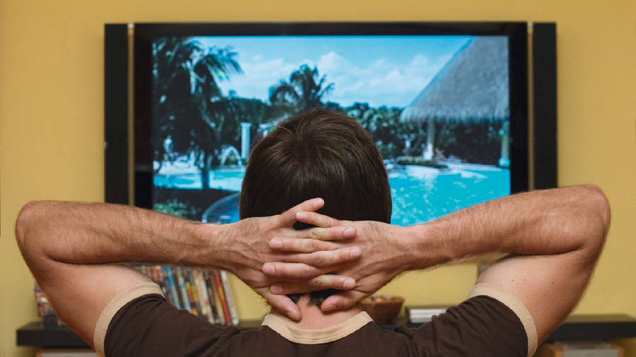 Person sitting back on a sofa watching TV with hands behind their head, looking satisfied